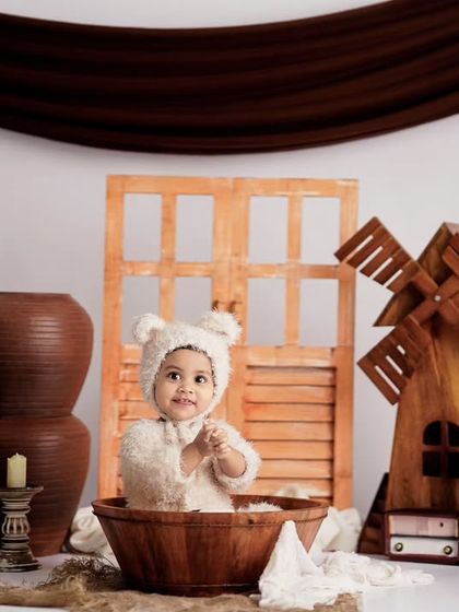 A cute toddler in a bear costume sits in a wooden bowl, part of a rustic studio scene with a windmill and terracotta pots.
