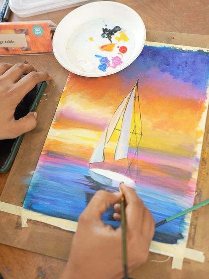 A close-up of a student working on a vibrant acrylic painting of a sailboat at sunset during a course session.