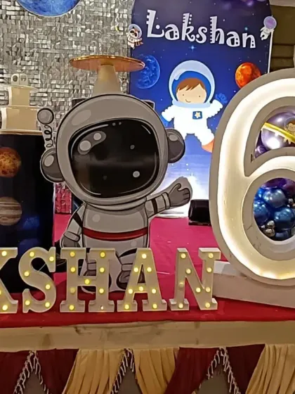 A close-up of the personalized details for Lakshan's space party. The setup includes a large astronaut cutout, a light-up number '6' with a planet inside, and illuminated letters for the name.