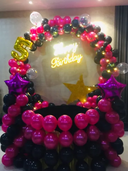 A chic birthday party photo booth created with a circular balloon frame in black and magenta. The setup is personalized with a number '5' foil balloon, star accents, and a 'Happy Birthday' neon sign.