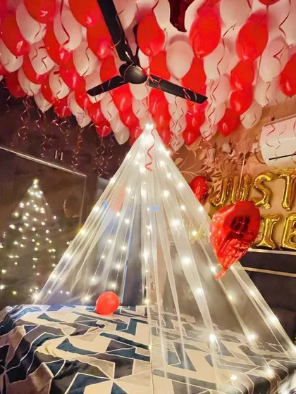 A cozy and romantic room decoration featuring a tent made of sheer fabric and fairy lights over the bed. The ceiling is filled with red and white balloons, and 'Just Married' foil letters are placed on the headboard.