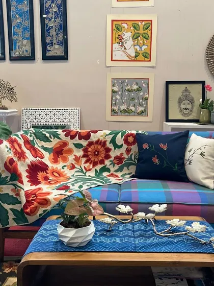 A cozy living room setup, featuring a colourful plaid sofa adorned with a floral throw, embroidered cushions, and a collection of Indian art on the wall.