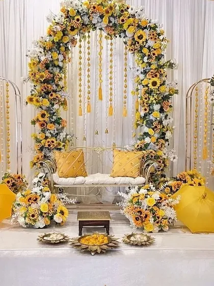 A complete Haldi ceremony stage, elegantly designed with a floral arch of sunflowers and white blossoms. The setup includes matching seating, decorative umbrellas, and traditional brassware for a cohesive look.