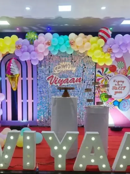A sweet and colorful candy-themed first birthday. The decor features a shimmering silver backdrop, pastel balloon arch with ice cream and donut foil balloons, and a large light-up number '1'.