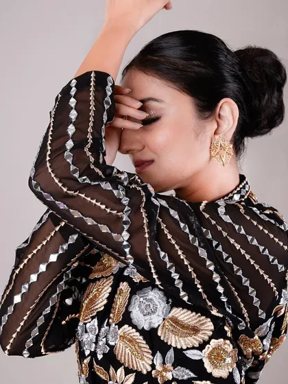 A close-up beauty shot highlighting the intricate gold and silver embroidery of a garment. The model's pose, partially covering her face, draws attention to the detailed craftsmanship and her elegant makeup.