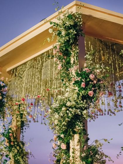 The floral-wrapped pillars of the Varmaala mandap against the twilight sky. The soft pastel colors of the roses and the delicate jasmine strings create a romantic and dreamy atmosphere.