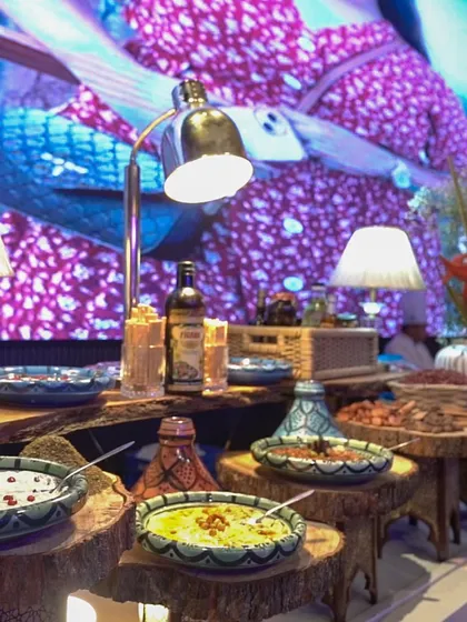 A rustic-chic display of Middle Eastern dips and salads, set on wooden logs and illuminated by elegant lamps, all against a backdrop of a live video wall.