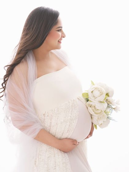 A radiant side-profile portrait against a bright, clean background. The simple styling with a white gown and bouquet puts all the focus on the joyful expression and beautiful baby bump.