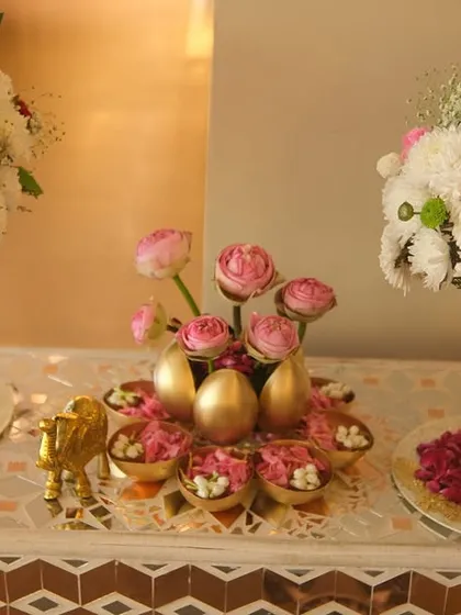 An elegant tabletop arrangement featuring large bouquets of white chrysanthemums and pink roses. The centerpiece includes golden eggs, small bowls of flowers, and a miniature golden elephant for a touch of opulence.