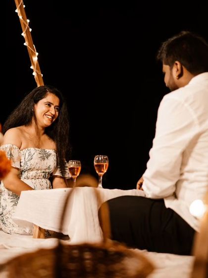 A candid shot of a couple interacting during their beach picnic at night. The image is framed by fairy lights, adding to the intimate and romantic atmosphere.