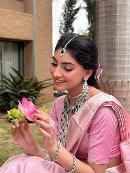 Holding a lotus, a symbol of purity and beauty. The makeup features soft pink tones on the eyes and cheeks, echoing the color of the flower and the saree.