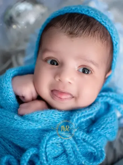 A sweet newborn wrapped in a bright blue knit blanket, peeking out with wide eyes and a tiny tongue sticking out.