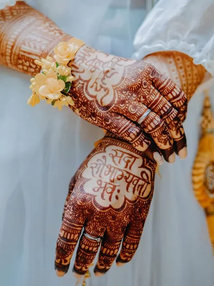 A detailed shot of the bride's hands during the Haldi ceremony, adorned with intricate henna and a delicate floral bracelet. The blessing 'Sada Saubhagyavati Bhava' is beautifully written.