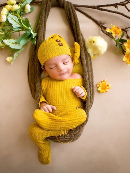 A newborn in a bright yellow outfit, sleeping in a hanging sling prop decorated with yellow flowers, creating a beautiful and artistic composition.