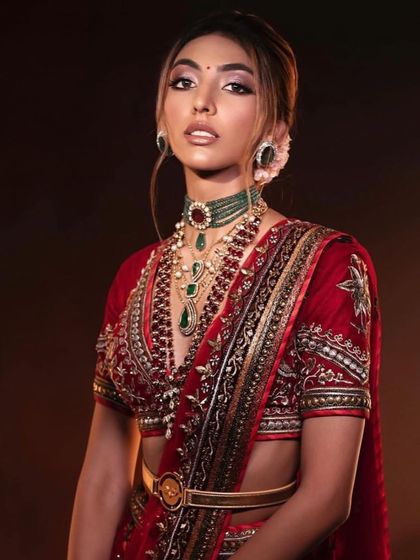 A regal bridal portrait. The makeup is soft yet defined, with a focus on the eyes, complementing the rich red and gold saree and the intricate jewellery.