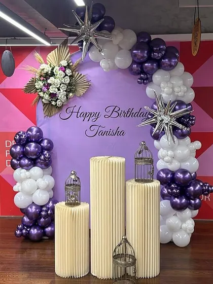 An elegant birthday backdrop in purple and white. This setup includes a personalized backdrop, a stylish balloon arch with floral and star accents, and decorative pedestals with birdcages.