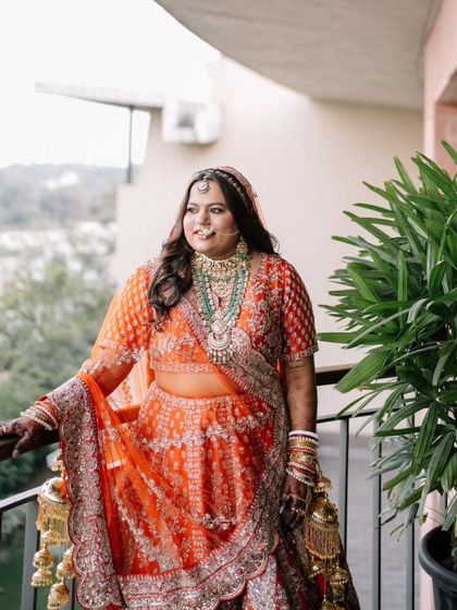 A beautiful shot of the bride on a balcony. Her orange and red lehenga is vibrant, and the makeup is designed to be bold yet elegant, perfect for a daytime wedding.
