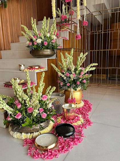 A beautiful arrangement at the base of a staircase, featuring large bouquets of tuberoses and roses in traditional brass pots (urlis). The floor is decorated with rangoli-style patterns made from flower petals.