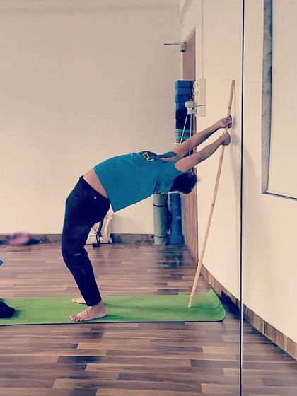 A participant uses the wall and a stick for a deep backbend. This supportive setup allows for a safe exploration of spinal extension, helping to increase flexibility and release tension in the back.