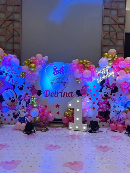 A grand Minnie Mouse themed first birthday for Delrina. The setup includes multiple backdrops, large balloon arches in pink and gold, and a light-up marquee number '1'.