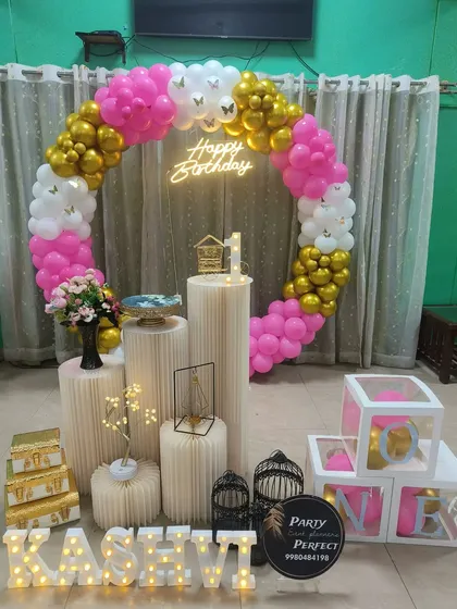 A bright and cheerful first birthday setup using a pink, white, and gold balloon ring, a neon sign, and personalized props for a complete look.