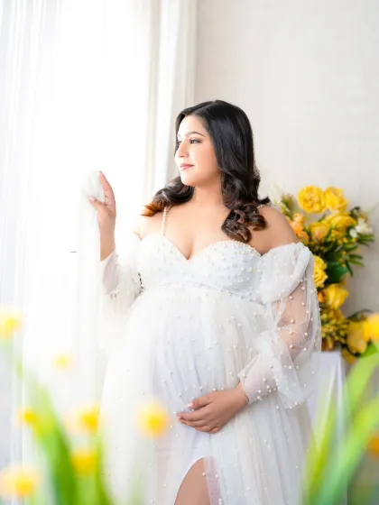 A touch of sunshine. The mother-to-be in a beautiful pearl-adorned white gown looks out a window, surrounded by bright yellow flowers that add a cheerful pop of color.