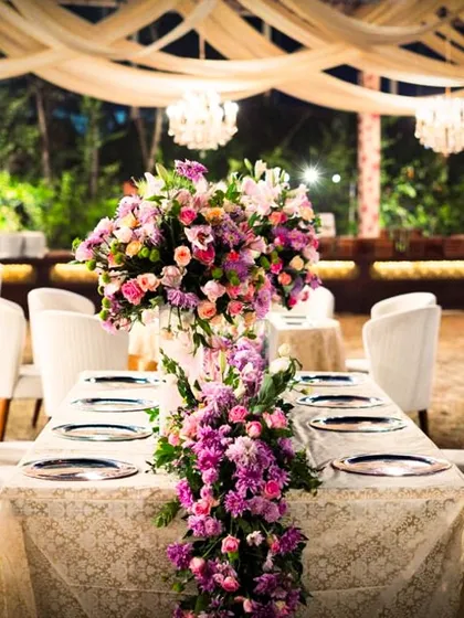 An elegant long table setup for a wedding reception, featuring a cascading floral runner in shades of purple and pink. This design creates a sense of intimacy and grandeur for guests.