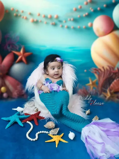 A fun and creative mermaid-themed photoshoot for a baby girl. The setup includes a knitted mermaid tail, seashell props, and an underwater-themed backdrop to create a magical scene.