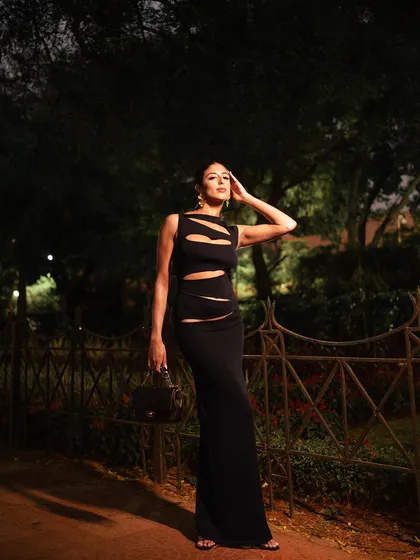 A full-body shot with a pensive, high-fashion pose. The ambient light of the park at night creates a moody and atmospheric backdrop for the sleek black dress.