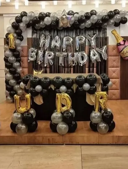 A stylish black and silver themed birthday stage for Rudra. The setup includes a balloon arch, 'Happy Birthday' foil letters, and the name spelled out with individual balloon columns.