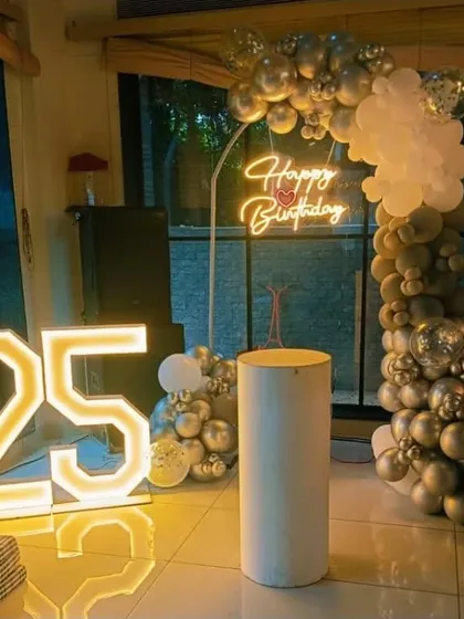 A 25th birthday party with a silver and white balloon arch, a 'Happy Birthday' neon sign, and a large '25' marquee light.