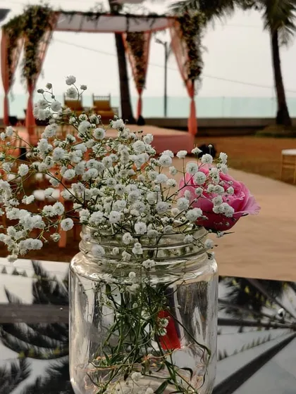 A close-up of a simple, rustic centerpiece. A mason jar with baby's breath and a single pink peony adds a touch of delicate charm to the guest tables.