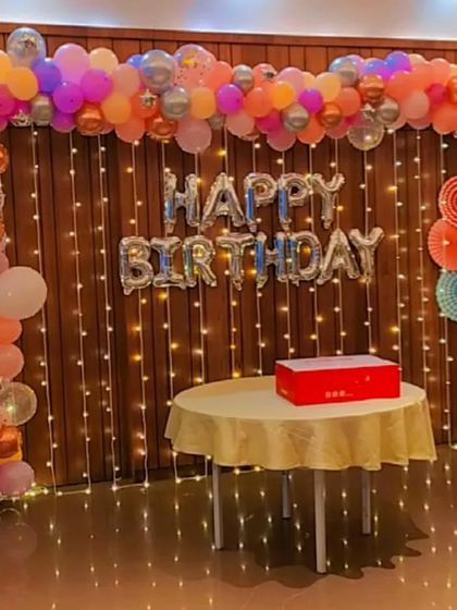This birthday decoration combines a colorful balloon garland with the warm glow of fairy lights against a wooden wall. The silver foil 'Happy Birthday' balloons add a festive touch, perfect for a cozy yet stylish party at home or in a hall.