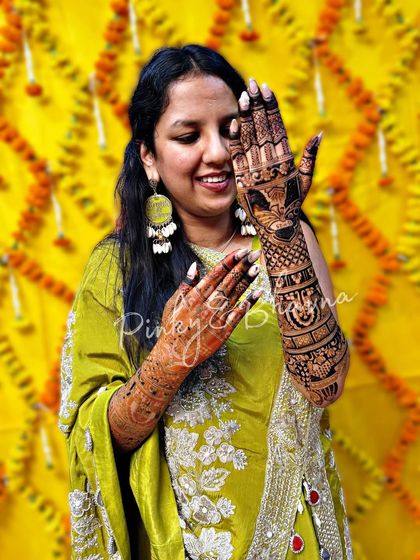 A happy bride poses against a vibrant yellow background, her detailed mehndi taking center stage. The design includes portraits and traditional Indian motifs, perfect for a grand wedding celebration.