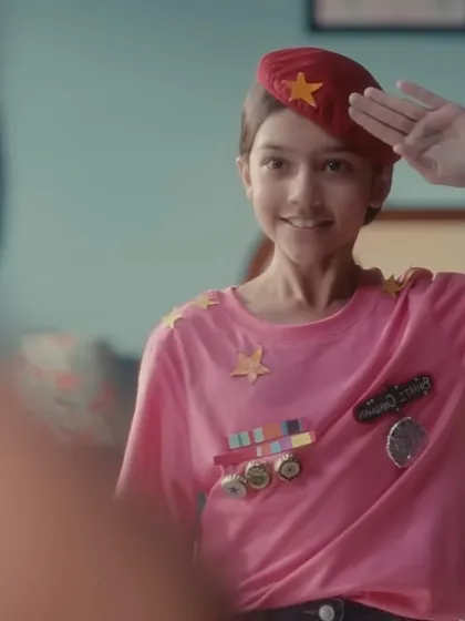 A close-up of the young character saluting her reflection. The combination of the red beret and the imaginative, self-made uniform was designed to show her early determination and the start of her journey.
