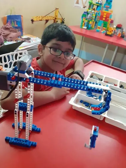 A smiling student proudly displays the complex crane mechanism he constructed using a VEX robotics kit.