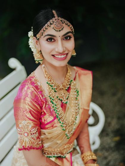 A smiling bride sits on a white bench, her beautiful jewelry and saree shining. This portrait captures the happiness and radiance of a bride on her wedding day.