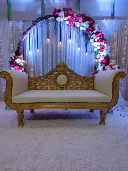 A classic and romantic baby shower stage. This design features a semi-circular floral arch, elegant hanging bulb lights, a traditional sofa, and a prop ladder spelling 'BABY' to complete the look.