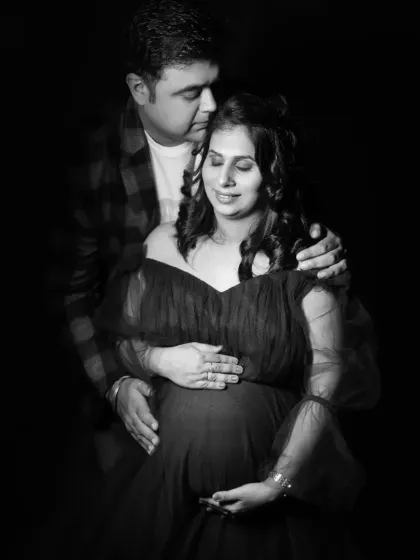 An intimate black and white studio portrait of a couple. The partner's hands gently rest on the mother-to-be's shoulders and bump.