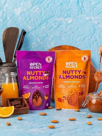 A styled group shot of two different almond snack flavors, Orange Cocoa and Caramel Sea-Salt. The composition uses fresh ingredients like orange slices and chocolate to hint at the flavors inside.