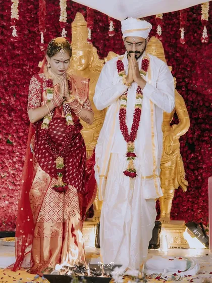 The bride and groom offering prayers together in front of a wall of red roses and golden deities.