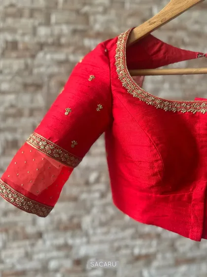 The quintessential red saree blouse, crafted in raw silk with a sheer panel on the sleeve and delicate hand embroidery along the neckline.