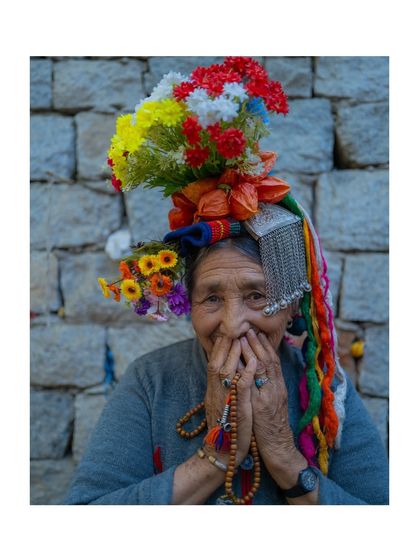 A shy, smiling portrait of a Brokpa woman from Ladakh. Her hands, holding prayer beads, are held to her face, drawing attention to her joyful expression and traditional headdress.
