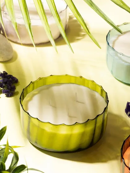 A fresh, summery shot of Lighthouse candles, styled with palm leaves and small flowers. The bright lighting and yellow background evoke a sense of sunshine and tropical scents.