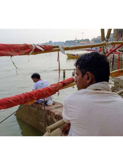 Two men sit by the edge of the Ganga in Varanasi, looking out at the water. The over-the-shoulder perspective creates an intimate and contemplative mood.