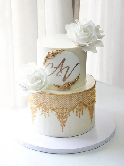 "Love layered in sweetness." A gorgeous, simple, and elegant two-tiered wedding cake featuring a white and gold design with lace stenciling and a custom monogram.