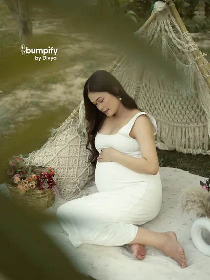 A peaceful moment captured during a boho-themed outdoor picnic. The mom-to-be, in a simple white dress, cradles her bump amidst macrame pillows and flowers.