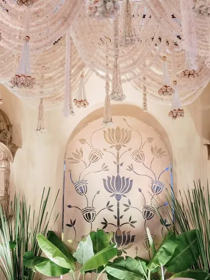 A detailed shot of the stage backdrop, where golden jharokhas and a lotus motif wall panel are framed by lush banana leaves and a canopy of white floral strings. This is a beautiful blend of tradition and nature.