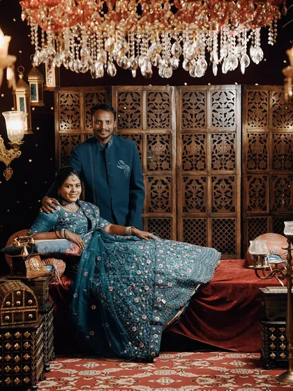 A regal pre-wedding setup with the couple in coordinated blue outfits. The intricate wooden screen, rich fabrics, and warm chandelier lighting create a royal ambiance.