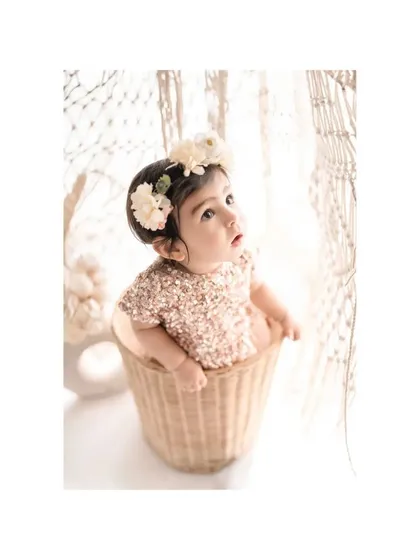 A baby girl in a sequined dress peeks out of a wicker basket, looking up with wonder. The clean, white background and bohemian-style props create a chic and adorable setup for a milestone photoshoot.